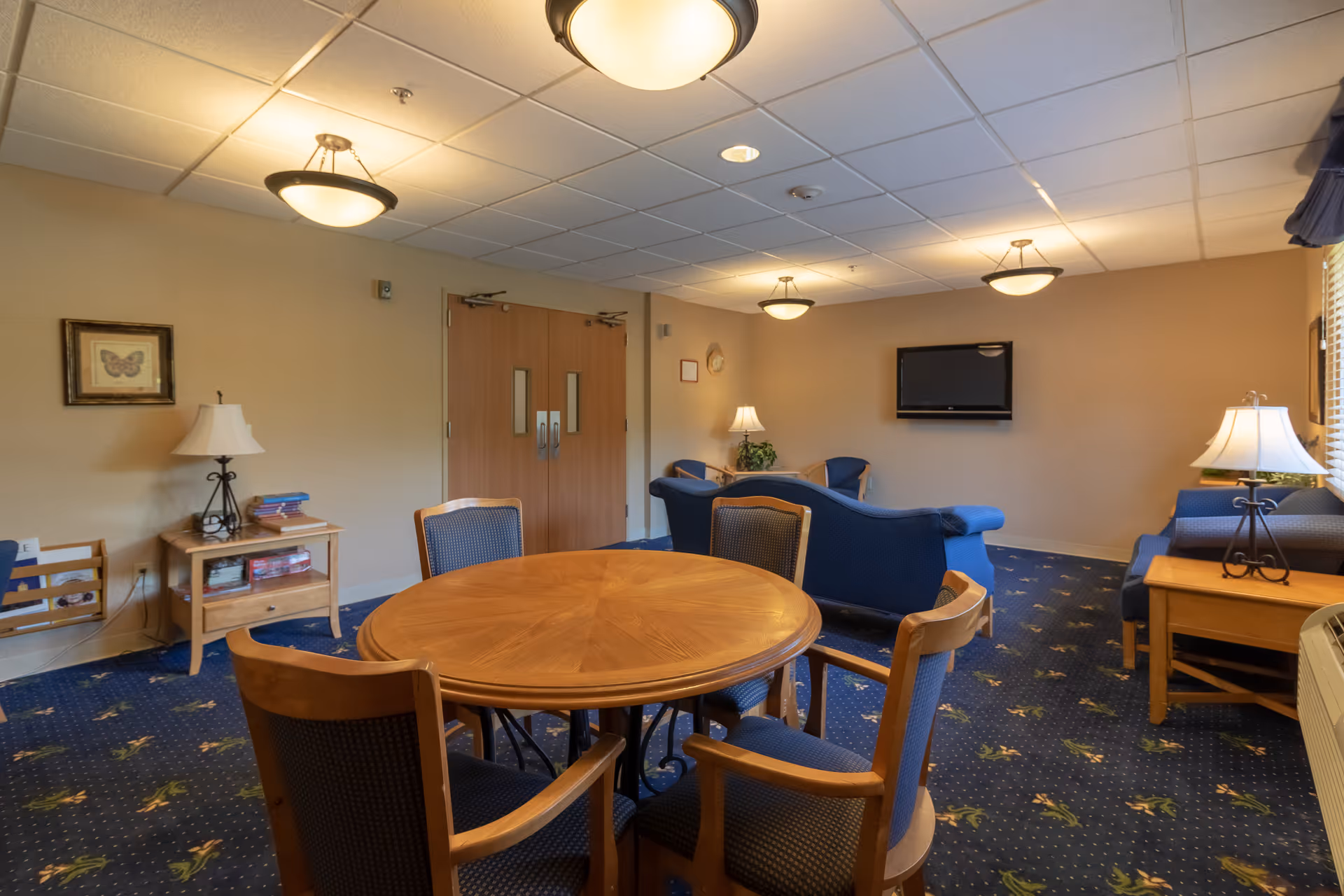 A senior living common room with a round wooden table and chairs, blue sofas, lamps, and a wall-mounted TV.