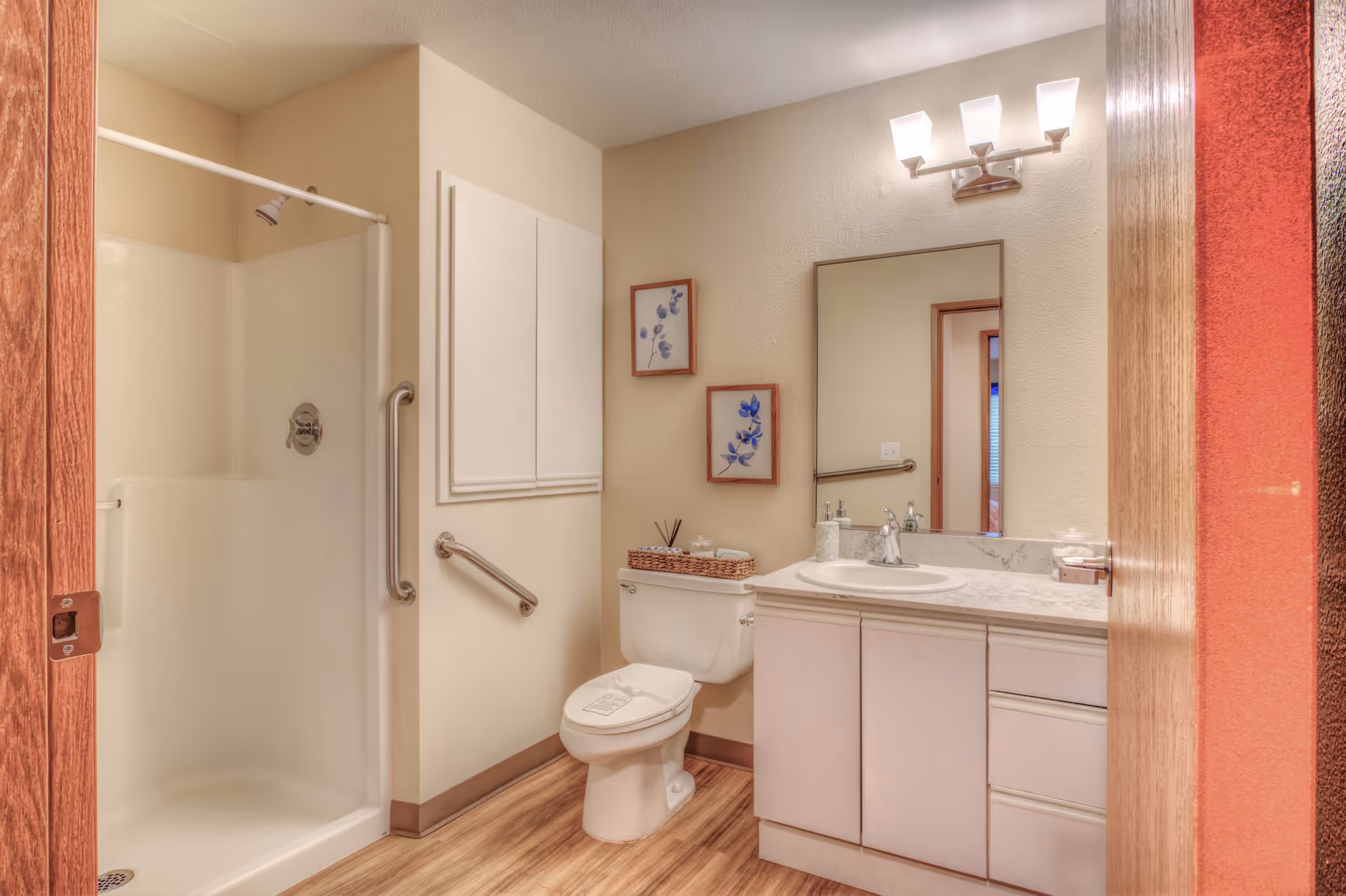 A clean and well-lit bathroom featuring a walk-in shower with grab bars, a toilet with a basket on top, a vanity with a sink and countertop, a large mirror above the sink, two framed floral pictures on the wall, and wooden flooring.