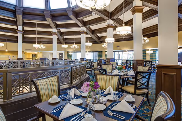 A spacious dining room with multiple tables set for a meal, featuring white napkins, plates, cups, and silverware. The room has high ceilings with wooden beams and chandeliers, and large windows allowing natural light to fill the space. The chairs have striped upholstery and the tables are decorated with floral centerpieces.