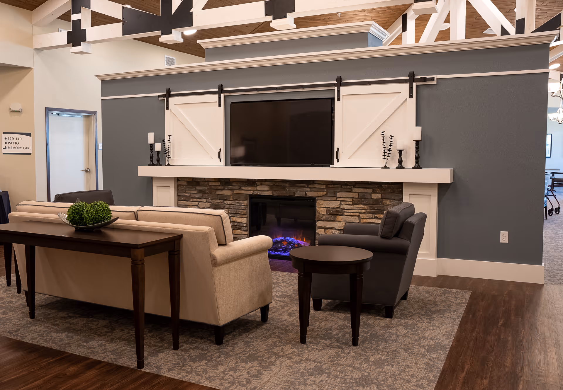 A cozy senior living common area featuring a beige sofa and a dark gray armchair arranged around a stone fireplace with a mounted TV above it. The fireplace has white wooden sliding barn doors on either side of the TV. The room has wooden flooring, a patterned area rug, and decorative candle holders on the mantel. A sign on the wall indicates directions to patio and memory care.