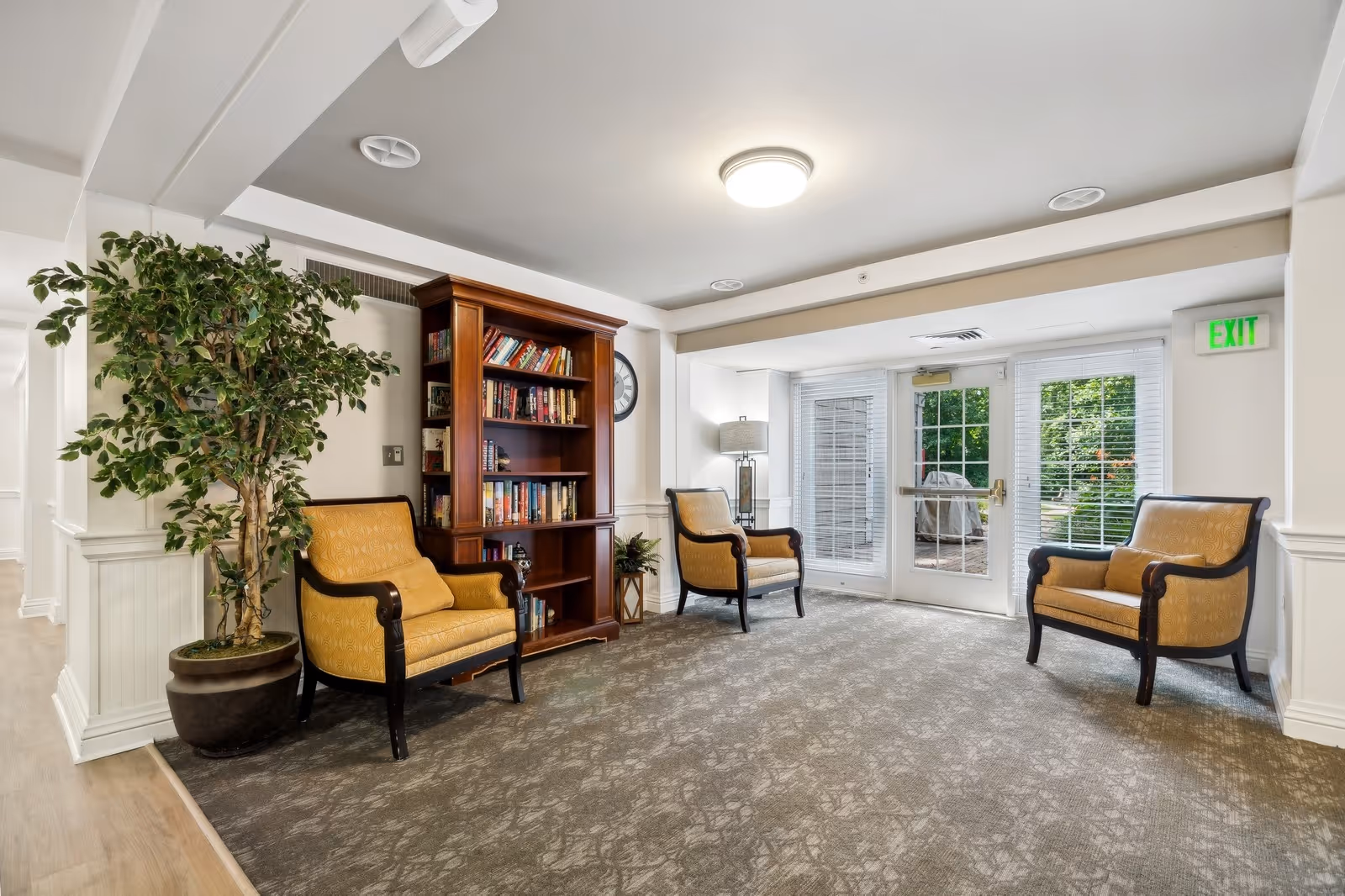 A cozy sitting area in a senior living facility with three yellow upholstered armchairs arranged around a wooden bookshelf filled with books. There is a large potted plant to the left and a floor lamp near the glass doors that lead outside. The room has carpeted flooring, white walls, and a ceiling light fixture.