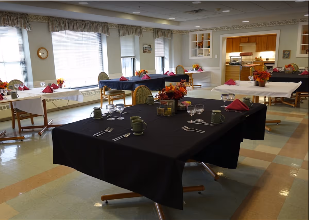 A dining room with several tables covered in black and white tablecloths, each set with green mugs, wine glasses, silverware, and red folded napkins. The tables have floral centerpieces with autumn-colored flowers. Large windows with blinds and valances allow natural light into the room. In the background, there is a kitchen area with wooden cabinets and a serving counter.