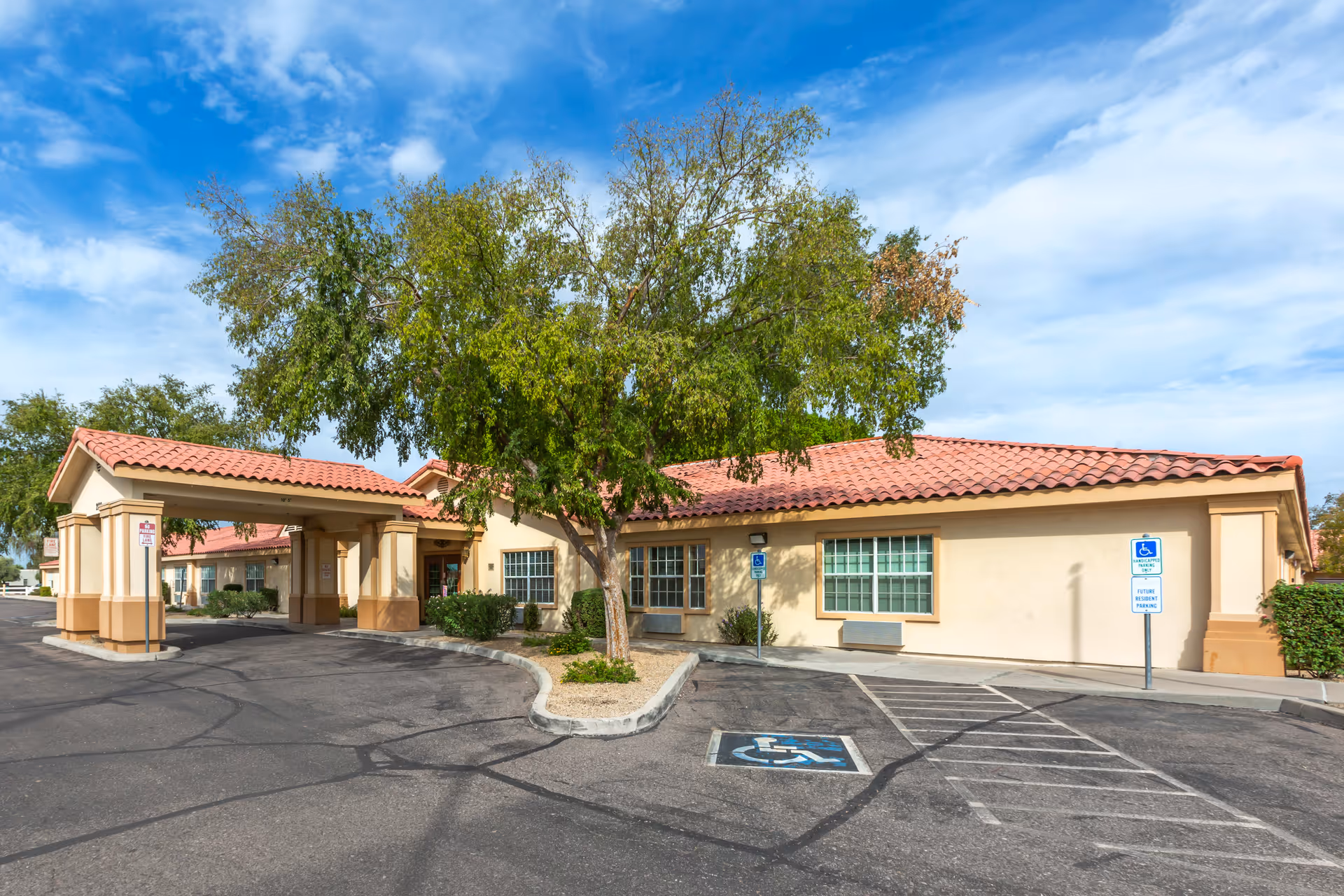 Exterior view of a single-story building with a red tiled roof and beige walls, featuring a covered entrance with columns. There is a tree in front of the building and a parking lot with marked handicapped parking spaces. The sky is partly cloudy.