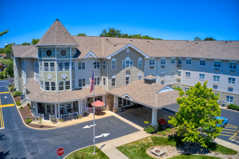 Exterior view of Addington Place of Collinsville, a large multi-story senior living facility with a turret-like architectural feature, multiple windows, a covered entrance, and a driveway with a stop sign and directional arrows. The building is surrounded by landscaping, including a tree and green grass, under a clear blue sky.