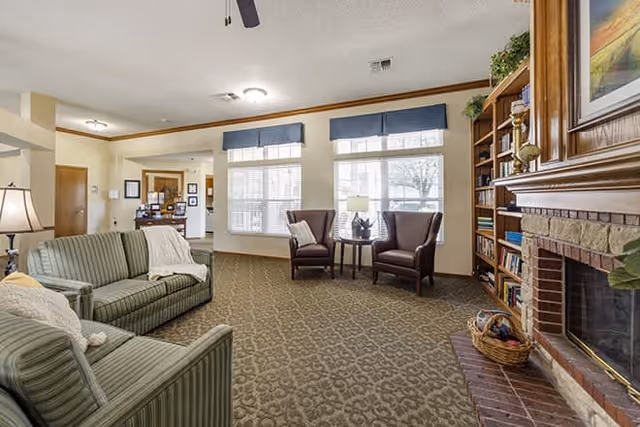 A cozy living room with patterned carpet, two green striped sofas, two brown armchairs with a small round table and lamp between them, large windows with blue valances, a wooden bookshelf filled with books and decorative items, and a brick fireplace with a wooden mantle.
