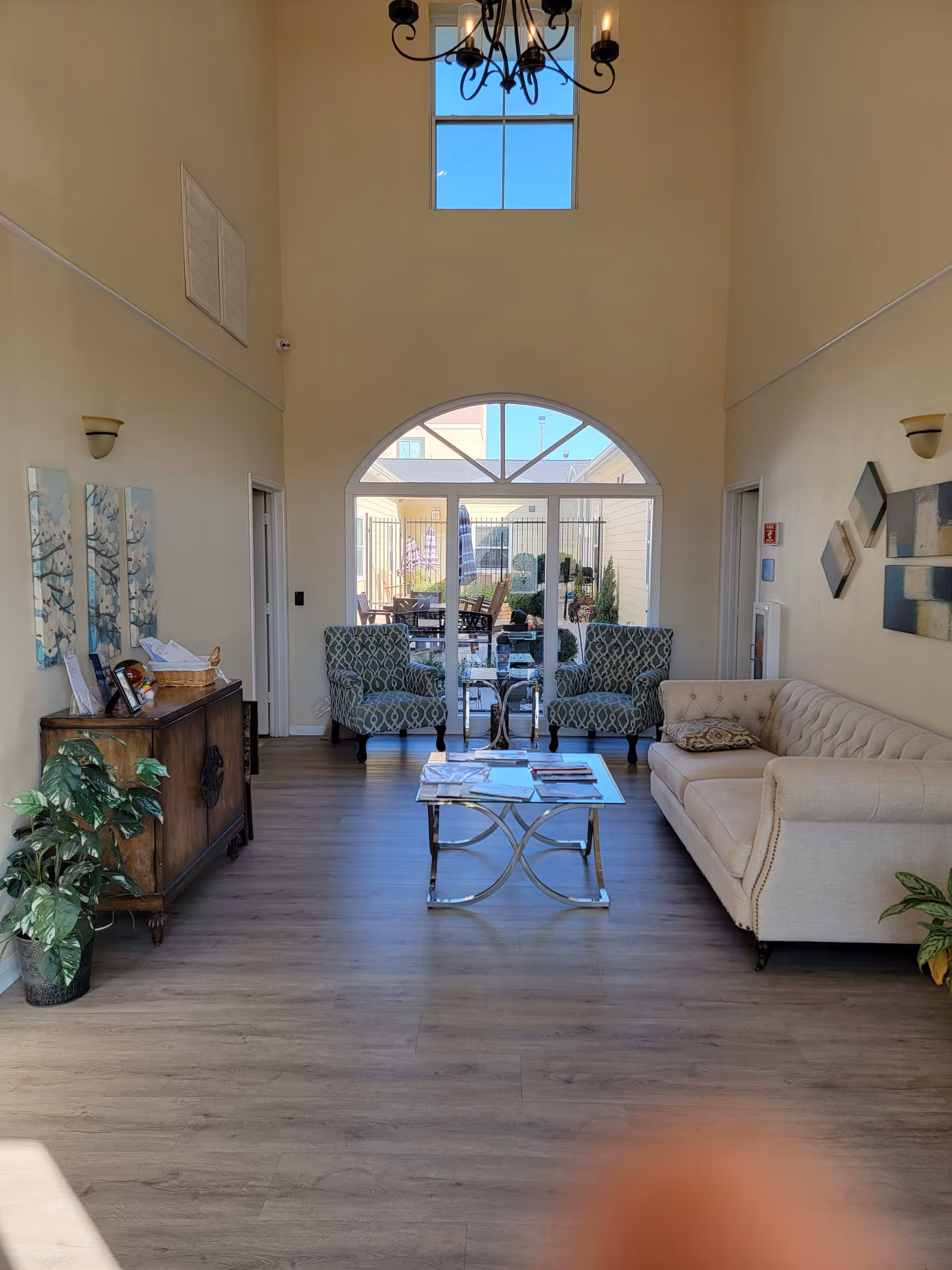 A bright and spacious living room area with high ceilings and a chandelier. The room features a beige tufted sofa on the right, two patterned armchairs facing the sofa, and a glass coffee table with magazines on it. There is a wooden cabinet with decorative items and a plant on the left side. Large glass doors with an arched window above lead to an outdoor patio area with tables and chairs. The walls are decorated with modern art pieces.