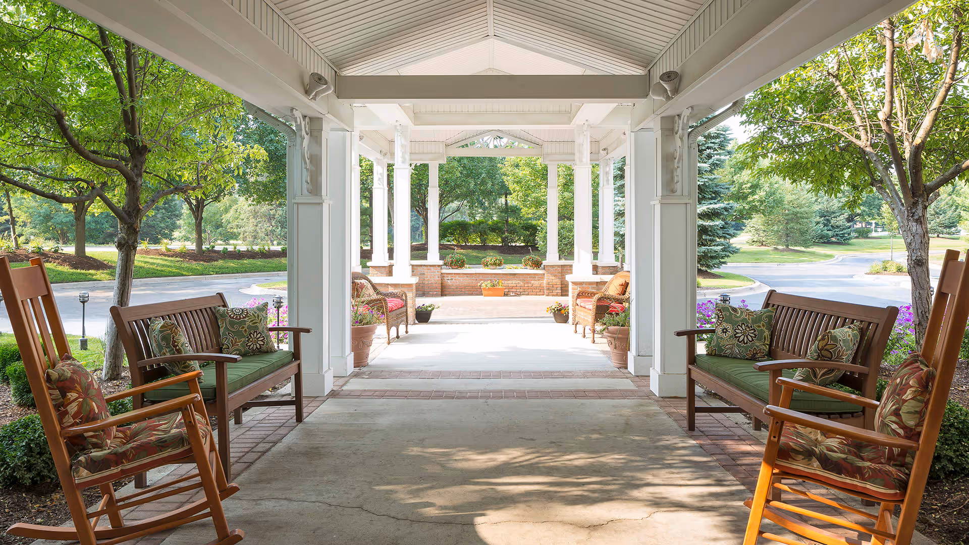 Covered outdoor seating area with wooden rocking chairs and benches with cushions, surrounded by trees and greenery in a garden setting.