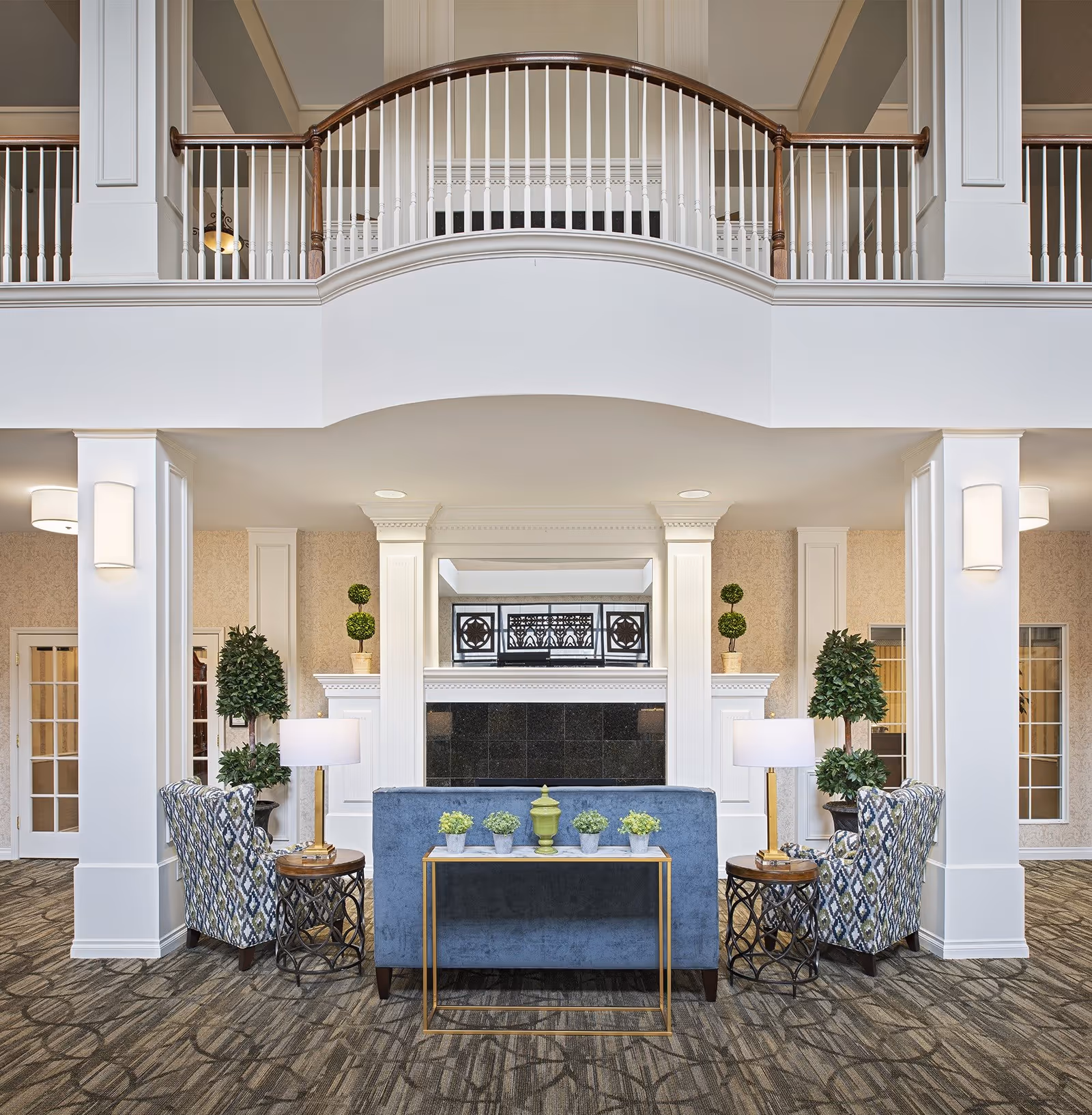 A spacious and elegant senior living facility common area with a high ceiling and a balcony railing above. The room features a blue sofa, two patterned armchairs, side tables with lamps, and a console table with small potted plants. There are decorative plants on either side of a fireplace with a black tile surround and white mantel. The walls are light-colored with subtle wallpaper, and the floor is carpeted with a patterned design.
