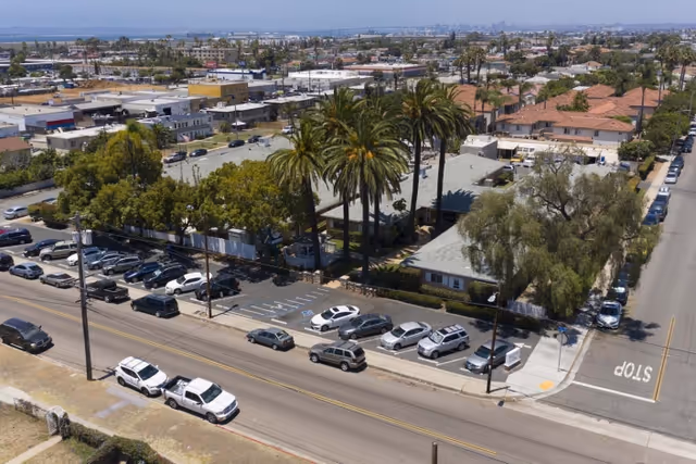 Aerial view of a residential neighborhood showing a single-story building surrounded by palm trees and other greenery, with a parking lot filled with cars in front. The area includes streets with parked vehicles and a mix of residential and commercial buildings in the background under a clear sky.