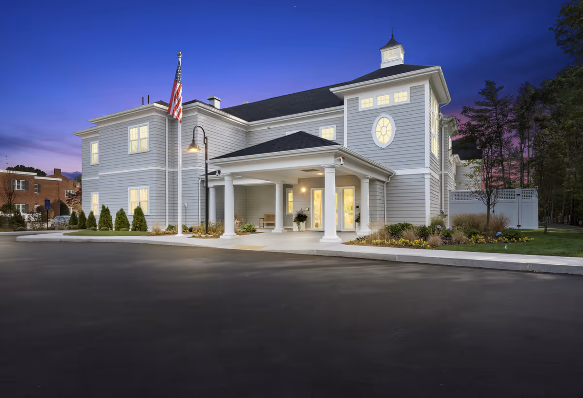 A large light-blue senior living facility with a covered entrance and American flag at dusk.