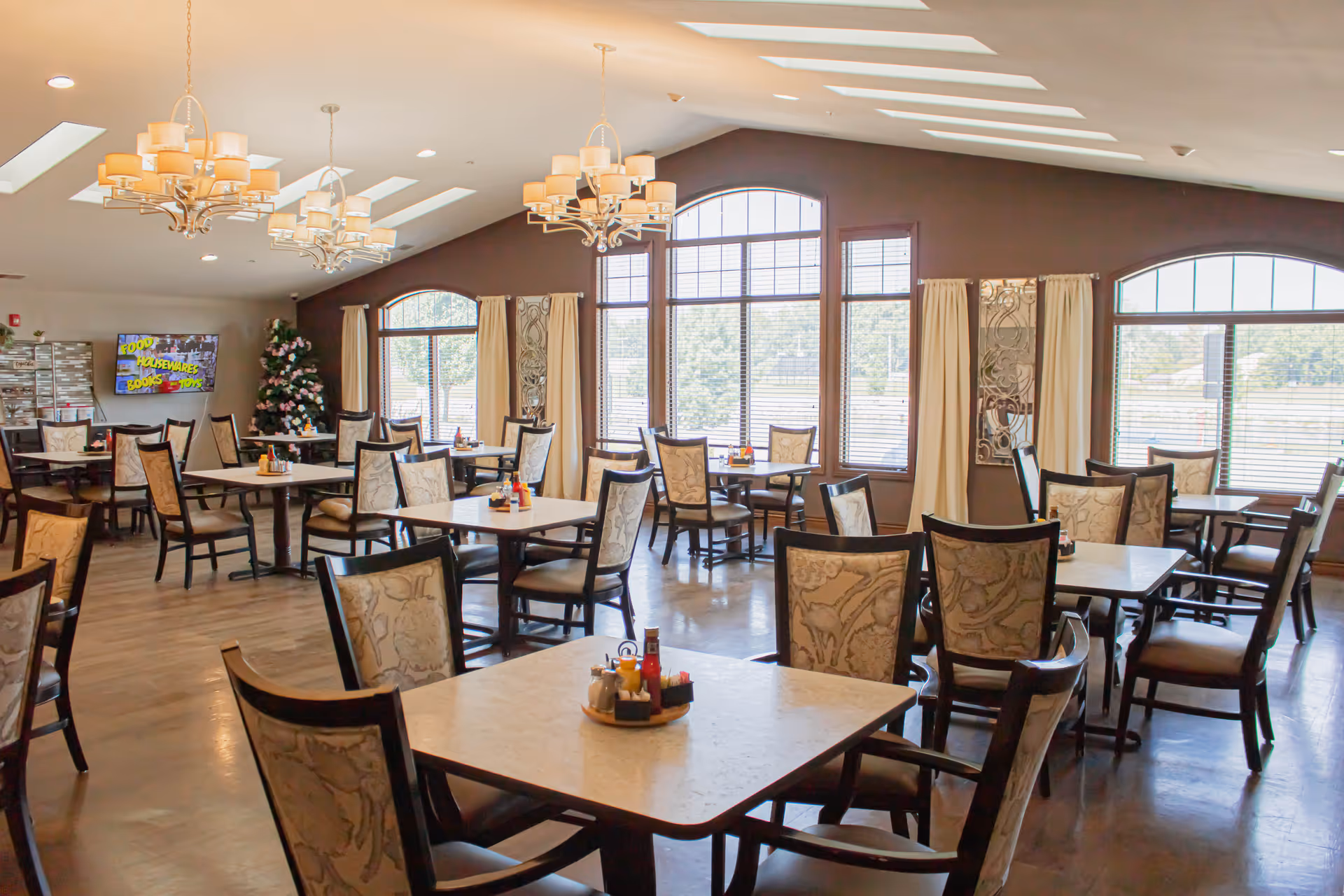 A spacious dining room with multiple square tables and cushioned chairs arranged neatly. The room has large windows with cream-colored curtains allowing natural light to fill the space. There are chandeliers hanging from the ceiling and a decorated Christmas tree in the corner. A television is mounted on the wall displaying colorful text.