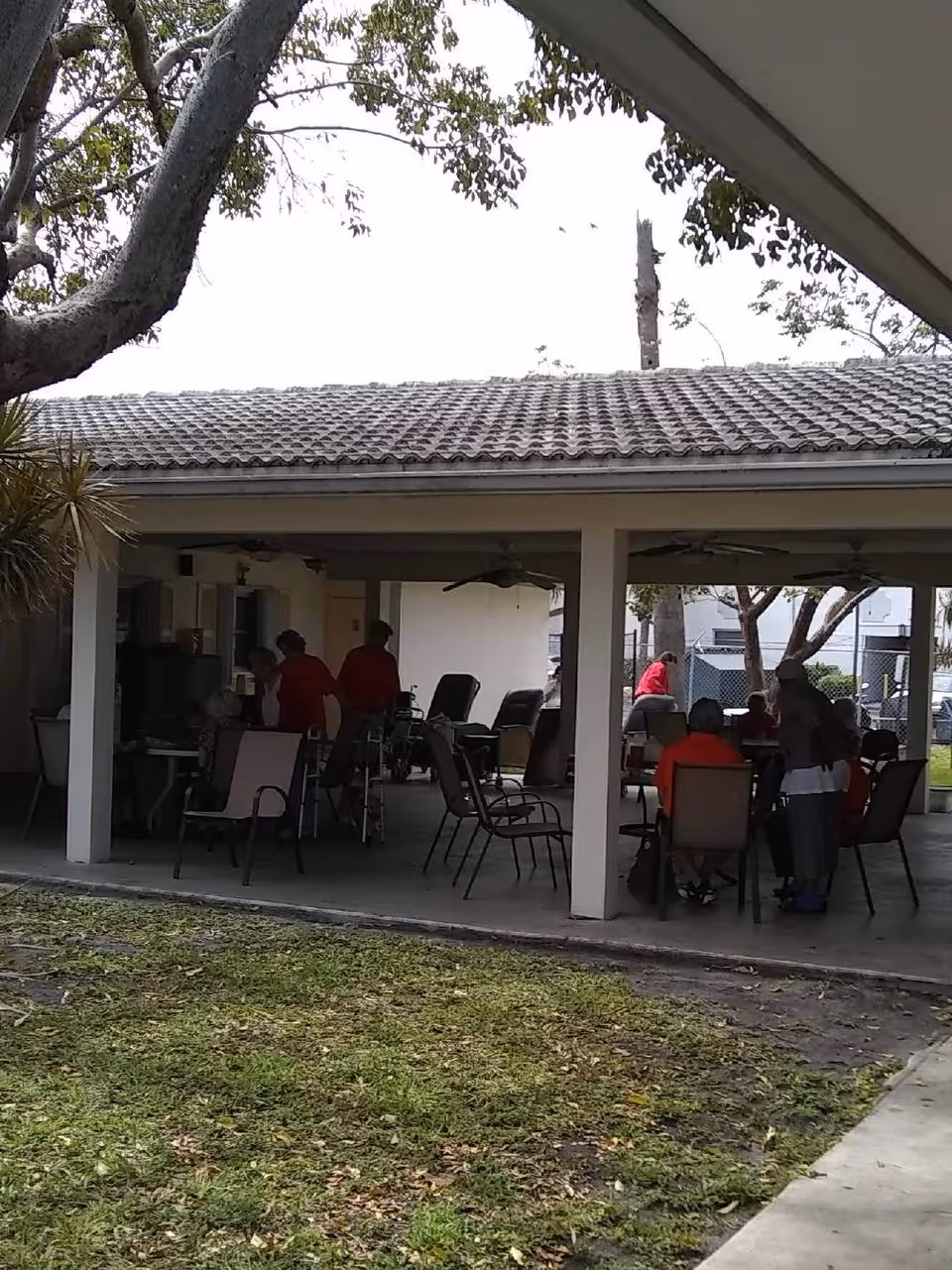 Outdoor covered patio area with several elderly people sitting and standing around tables and chairs, some in wheelchairs. The area is shaded by a tiled roof supported by white pillars, with trees and grass visible in the foreground.