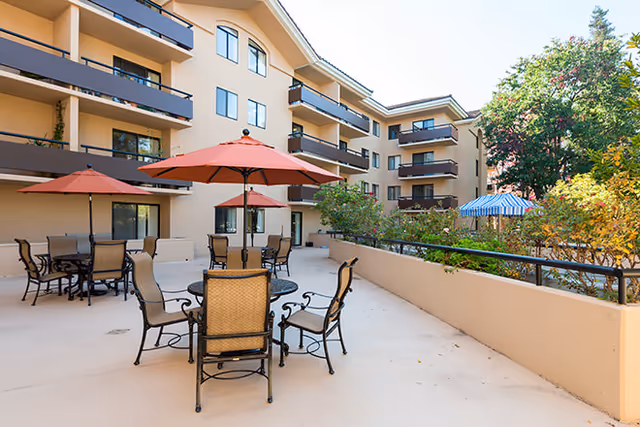 Outdoor patio area at Brookdale Redwood City with multiple tables and chairs under red and blue umbrellas, surrounded by a beige multi-story building and greenery.