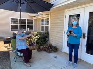 An outdoor patio area at Marbella Marysville with two elderly women wearing masks. One woman is seated on a chair under a large umbrella, holding a phone or camera, while the other woman stands near a glass door holding a camera. There are potted plants and bushes near the building's beige siding.