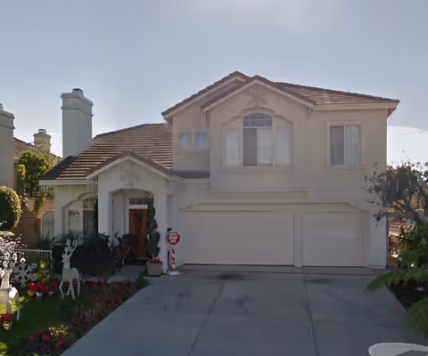 Front exterior view of a two-story residential home with a beige facade, a tiled roof, and a three-car garage. The driveway is concrete, and there are decorative plants and holiday decorations including reindeer and a stop sign near the entrance.