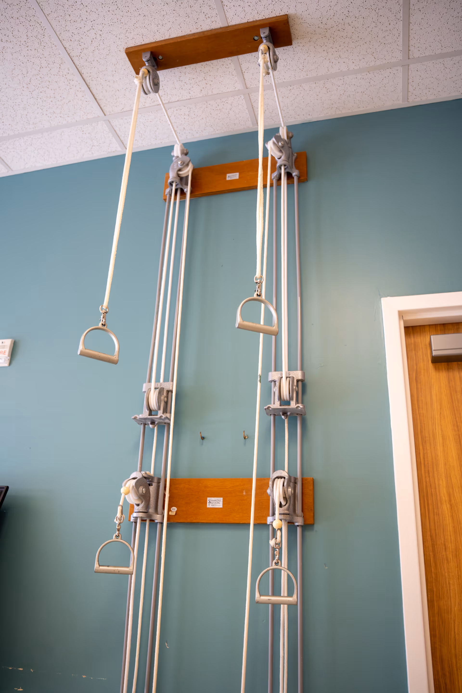 Wall-mounted rehabilitation pulley system with ropes and hand grips against a teal wall in a therapy room.
