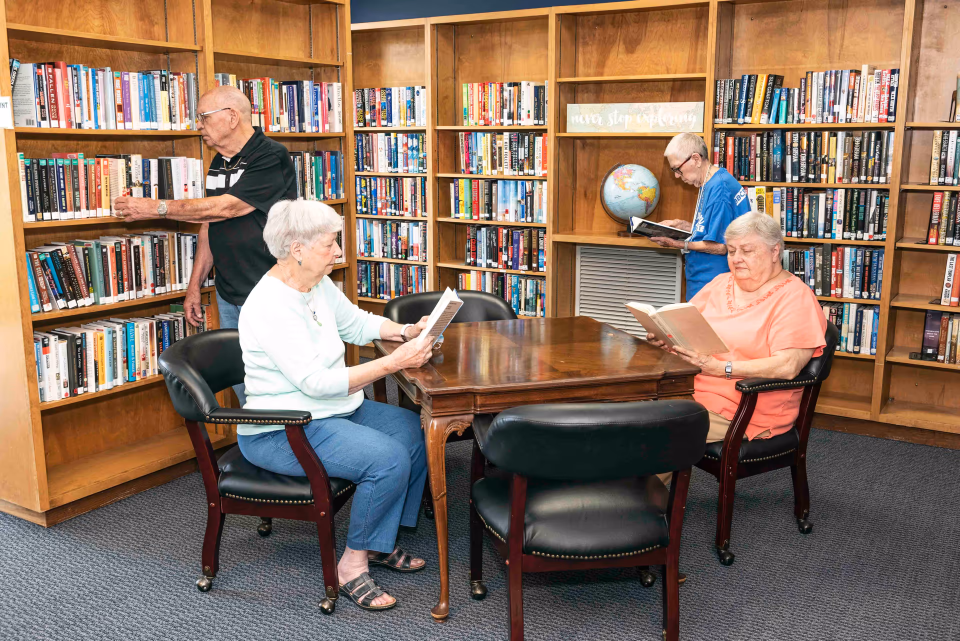 Four elderly individuals in a library room with wooden bookshelves filled with books. Two women are seated at a wooden table reading books, while two men are standing near the bookshelves also reading. A globe is visible on a shelf in the background.