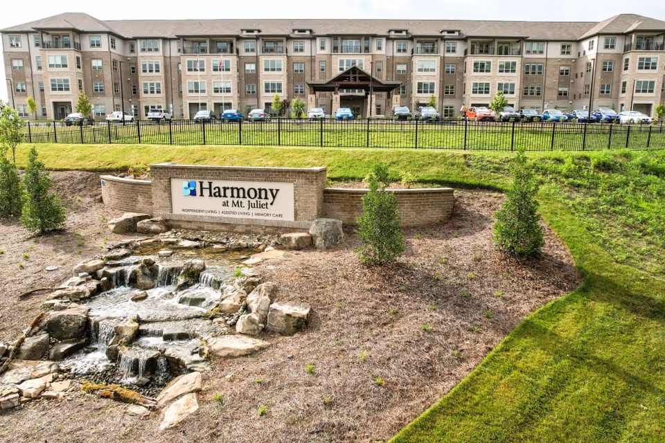Exterior view of Harmony at Mt. Juliet senior living facility showing a multi-story building with numerous windows and a covered entrance. In the foreground, there is a landscaped area with a small cascading water feature made of rocks, some small bushes, and a sign displaying the facility's name and services. Several cars are parked in front of the building.