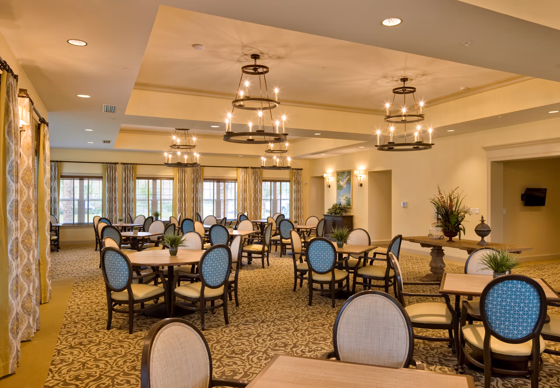 Spacious, well-lit communal dining room with round tables and upholstered chairs beneath chandelier lighting.