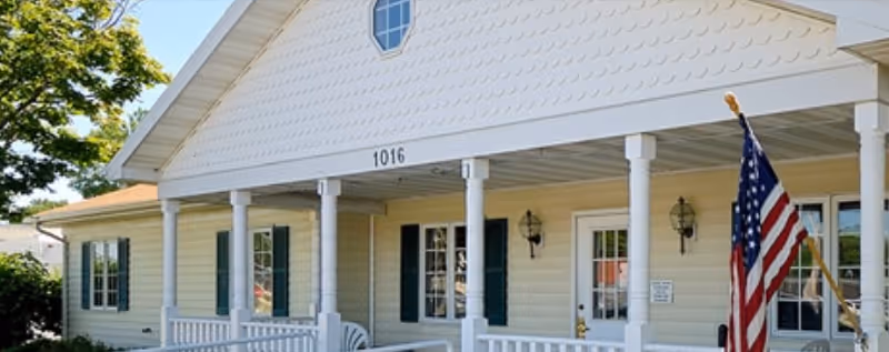 Beige single-story memory care facility with a covered front porch and American flag.