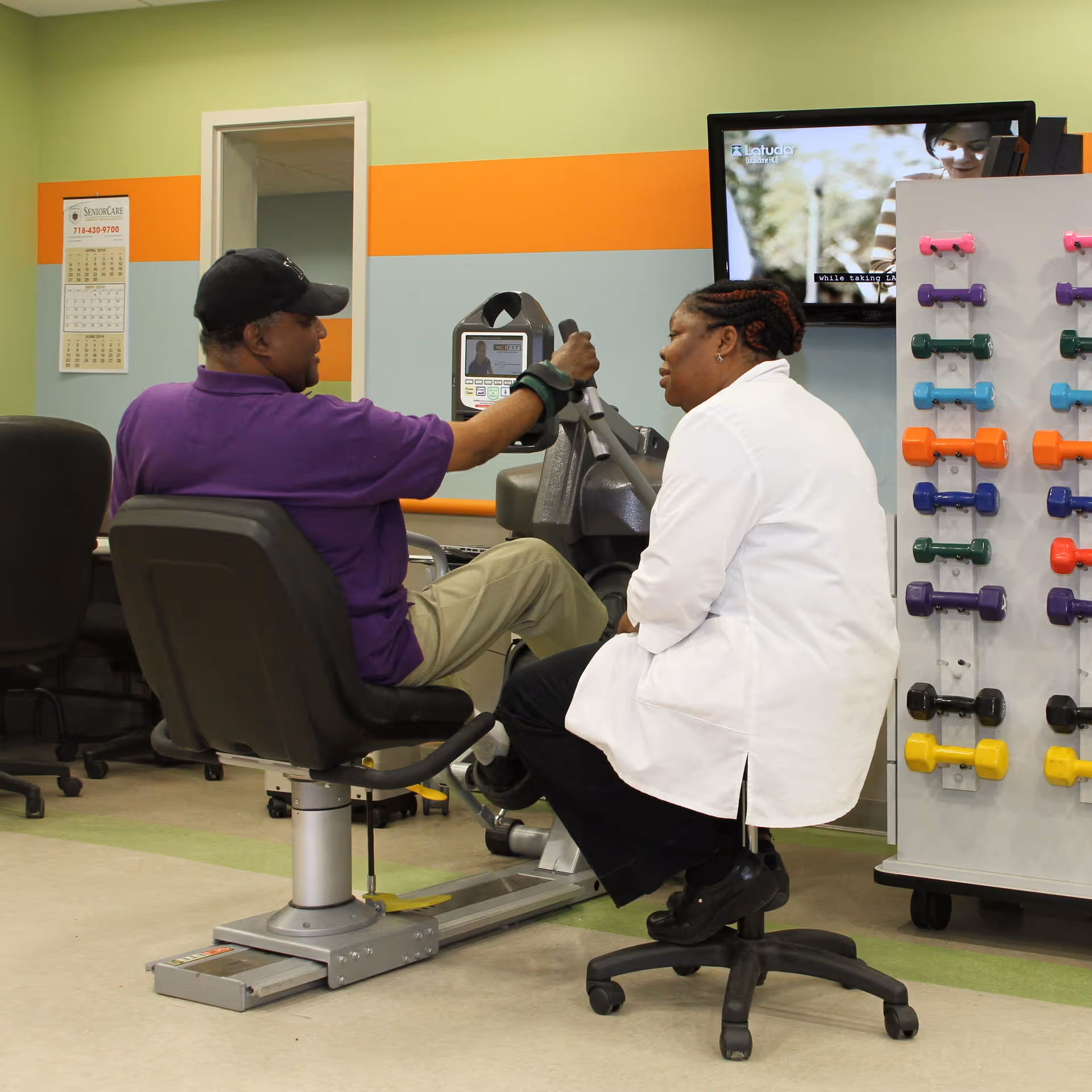 A man in a purple shirt and black cap is using a seated exercise machine while a woman in a white coat sits on a rolling stool beside him, providing assistance. The room has colorful walls with orange and blue stripes, a rack of colorful dumbbells, and a TV mounted on the wall.