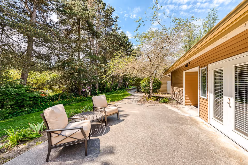 Outdoor patio area with two cushioned chairs and a small round table on a concrete surface next to a brown building with white-framed glass doors. The patio overlooks a green lawn and trees under a partly cloudy blue sky.