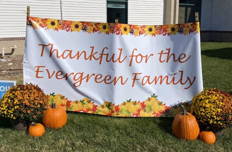 A large white banner with autumn-themed decorations including sunflowers and pumpkins. The banner reads 'Thankful for the Evergreen Family' and is displayed outdoors on a grassy area in front of a building. There are potted chrysanthemums and several pumpkins placed on the grass in front of the banner.
