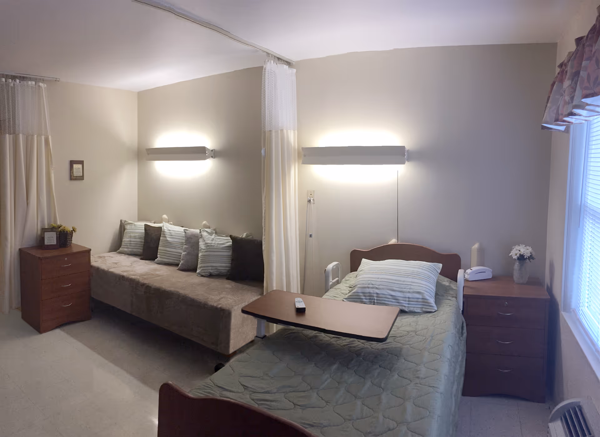 A senior living bedroom with a single hospital-style bed and overbed table, a daybed with cushions, bedside dressers and soft wall lighting.