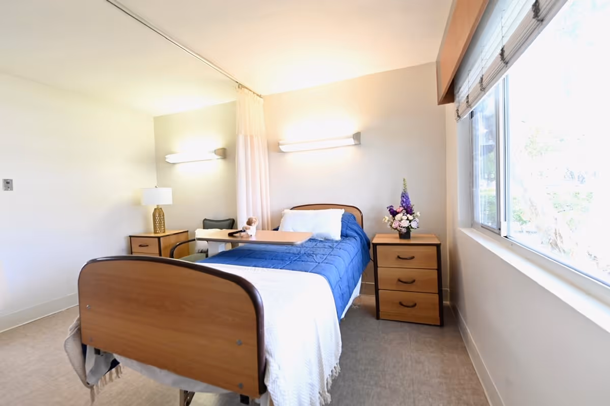 A bright and clean bedroom in a healthcare facility featuring a single bed with a blue blanket and white pillow. There is a wooden bedside table with a vase of flowers on the right side of the bed, and another small table with a lamp and chair on the left side. A large window on the right wall lets in natural light, and the room has neutral-colored walls and flooring.