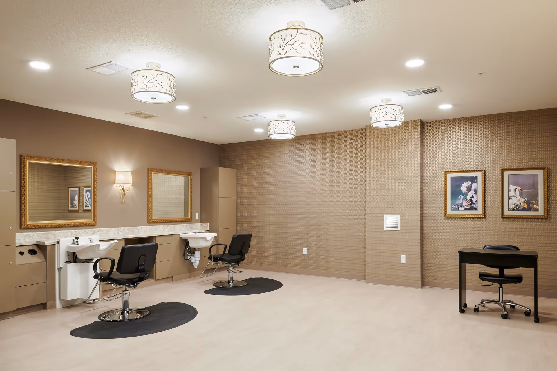 A spacious salon area with two black salon chairs in front of white sinks and large gold-framed mirrors mounted on a beige wall. The room has light-colored flooring, patterned wallpaper on one wall, two framed floral paintings, a small black desk with a black office chair, and decorative ceiling lights.
