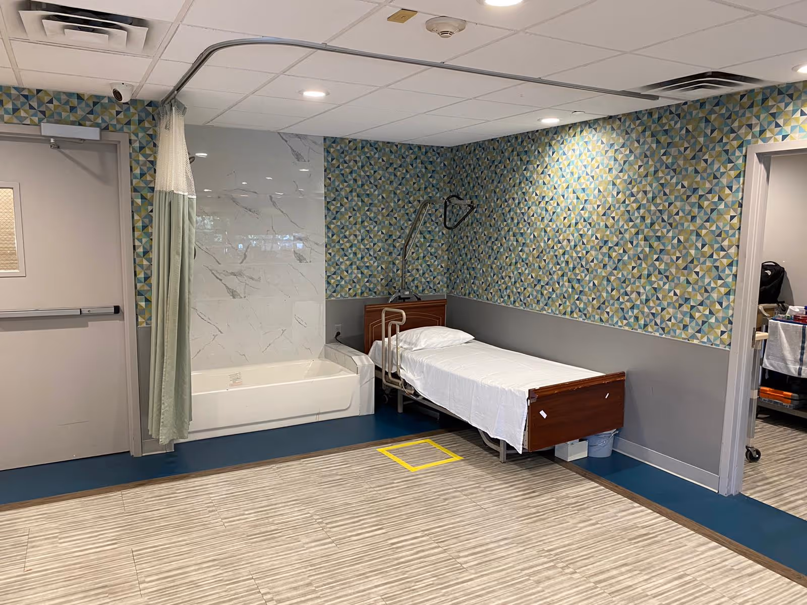 A room in a care facility featuring a single hospital-style bed with white sheets next to a bathtub with a shower curtain. The walls have a geometric patterned wallpaper above a gray lower section. The floor has a combination of blue and beige tiles, and there is a door on the left side and an open doorway on the right side leading to another room.