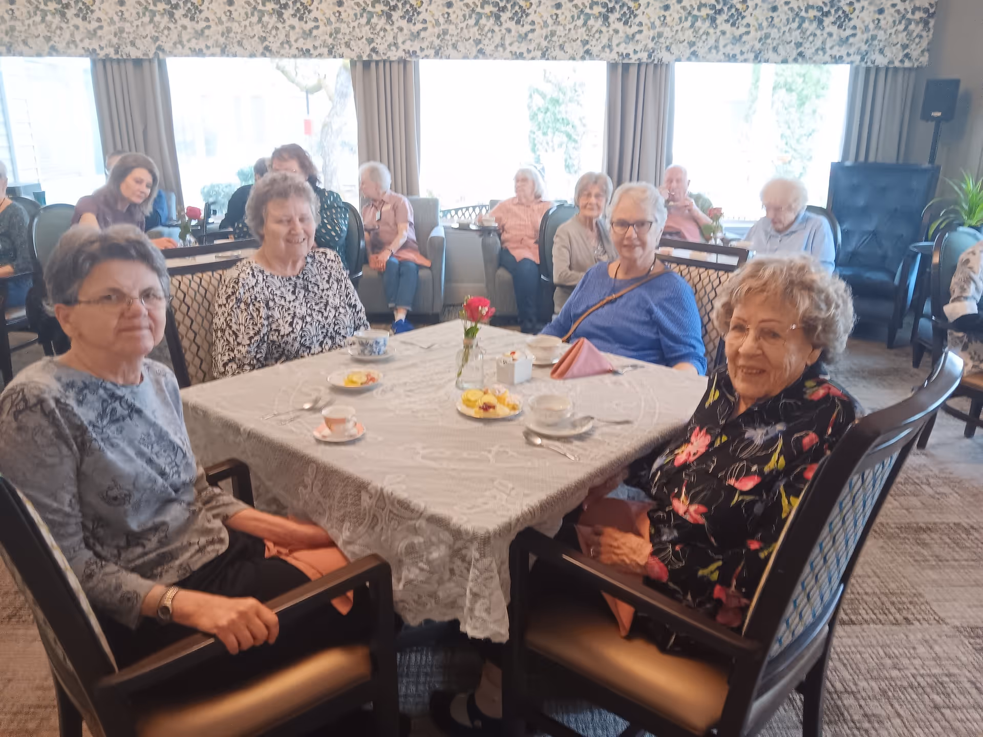A group of elderly women sitting around a table covered with a lace tablecloth in a well-lit room. The table has cups, plates with small snacks, and a vase with a red flower. More elderly people are seated in the background near large windows with curtains.