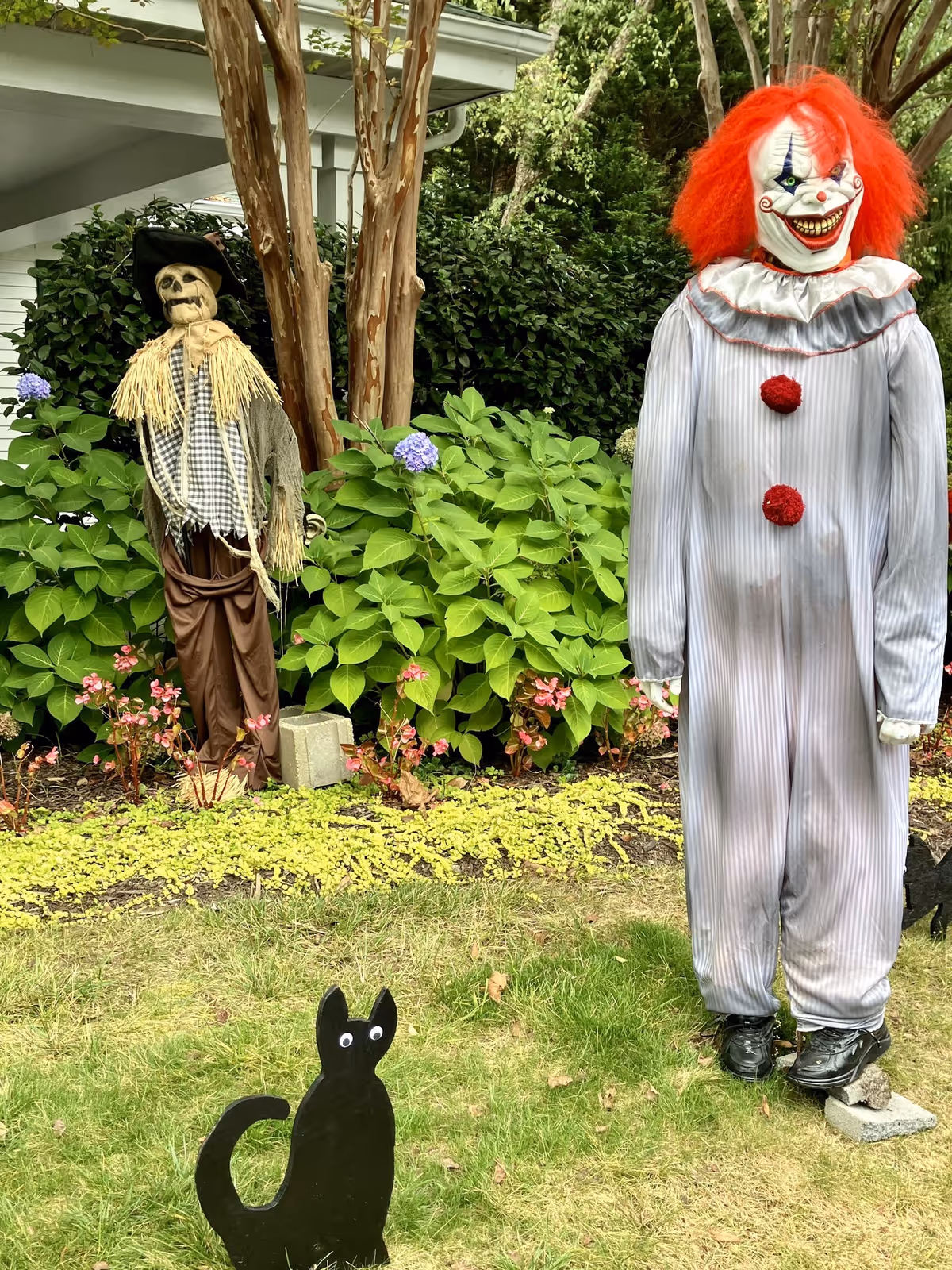 Outdoor garden area with Halloween decorations including a scary clown figure with bright red hair and a skeleton scarecrow dressed in rustic clothing, surrounded by green bushes and flowers. A black cutout of a cat with googly eyes is placed on the grass in the foreground.