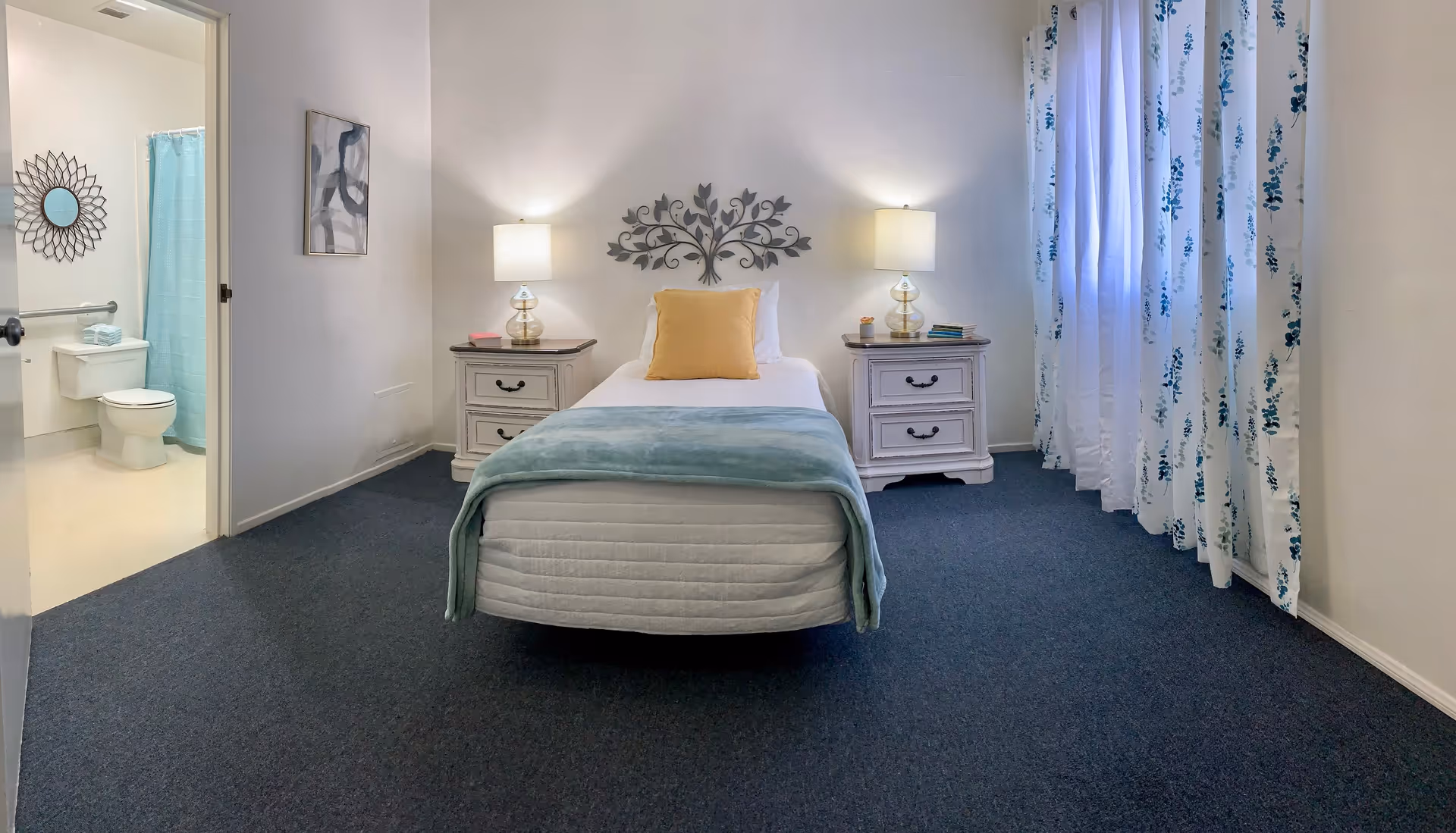 A neatly arranged bedroom with a single bed centered against the wall, flanked by two white nightstands each with a lamp. The bed has a yellow pillow and a light blue blanket. To the left, an open door reveals a bathroom with a toilet, a blue shower curtain, and a decorative mirror on the wall. On the right side, there is a window with white and blue floral curtains.