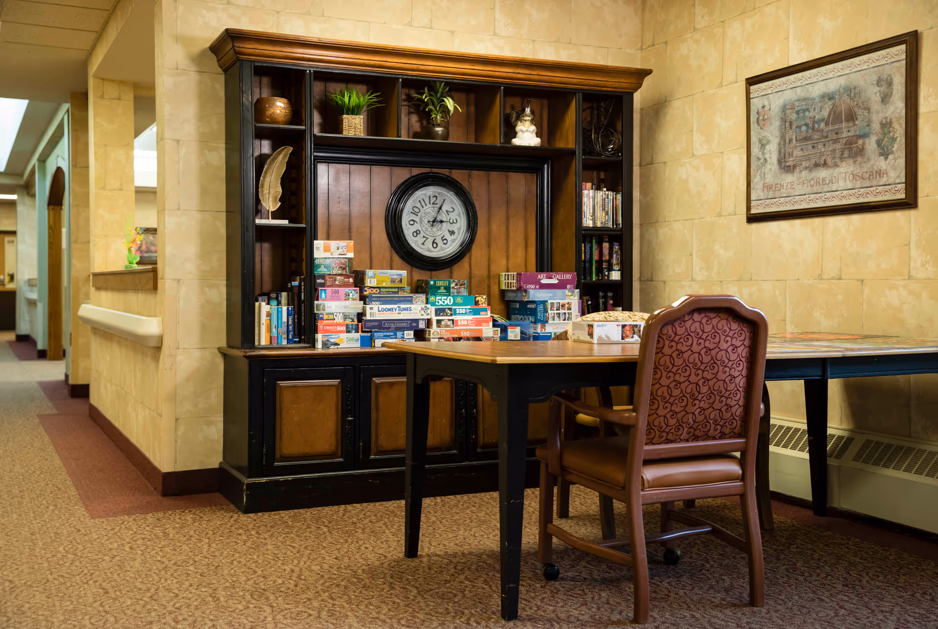 A cozy interior space featuring a wooden table with a cushioned chair in front of a large dark wood shelving unit. The shelves hold various board games and puzzles, along with decorative plants and a large round clock. The walls are tiled in a beige color, and a framed artwork hangs on the right wall. The carpet is patterned, and a hallway is visible to the left.