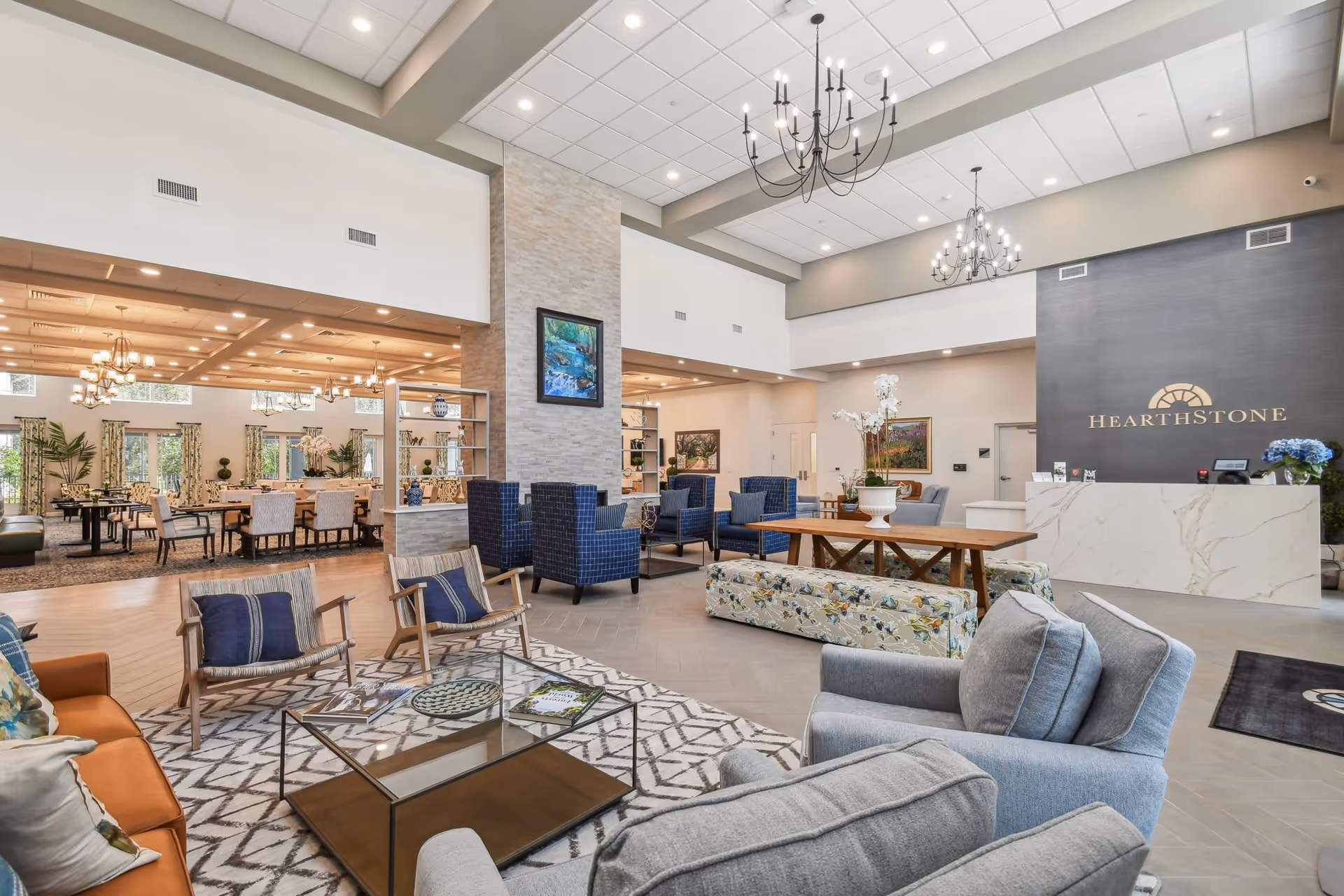 Spacious and well-lit senior living facility common area with modern furniture including armchairs, sofas, and a glass coffee table. The room features high ceilings with chandeliers, a reception desk with the HearthStone logo on the wall behind it, and a dining area with tables and chairs visible in the background. Decorative plants, paintings, and a patterned rug add to the welcoming atmosphere.