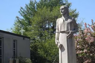 Outdoor statue of a standing figure beside a building with trees and blossoms