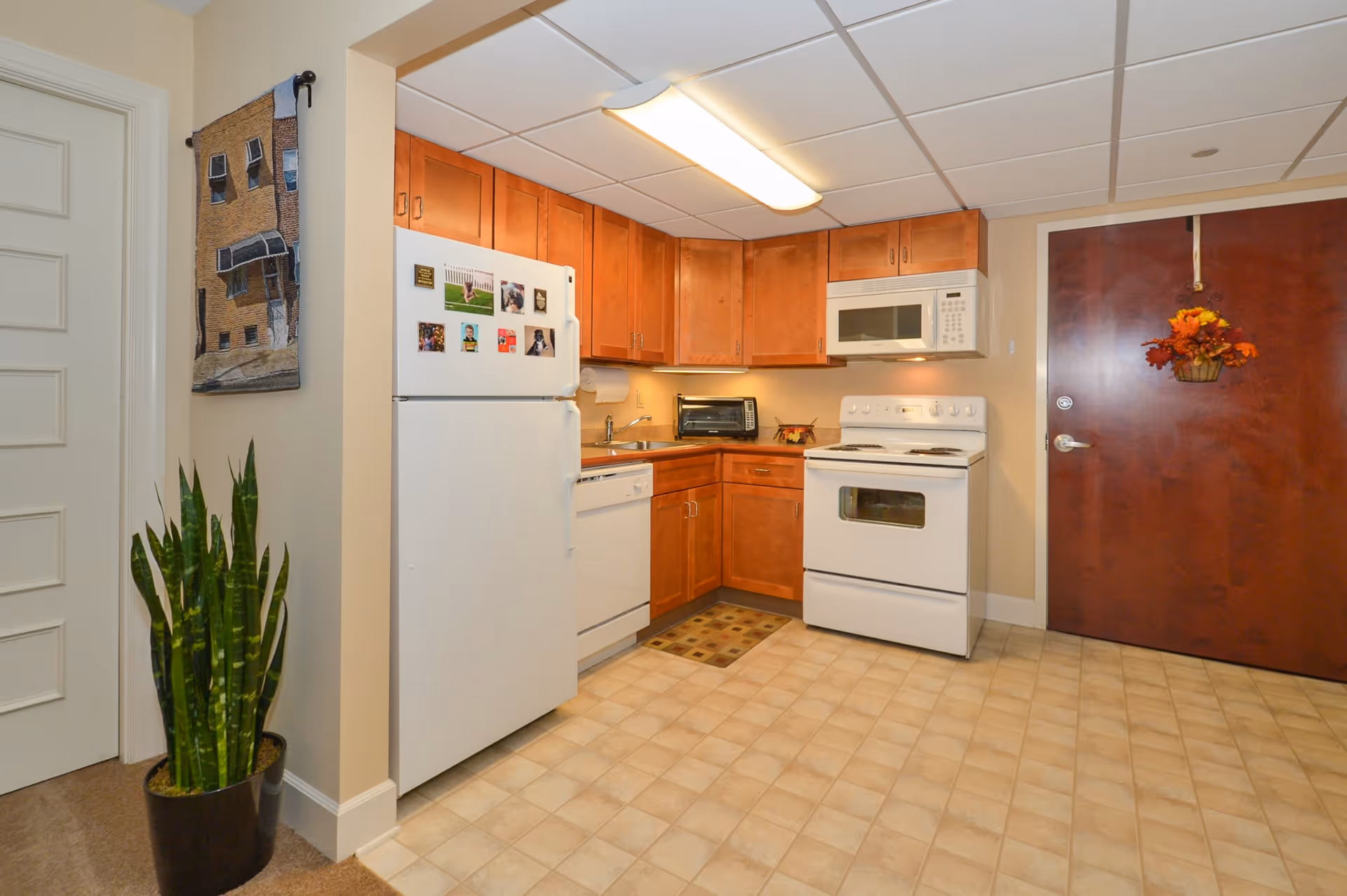 A small kitchen area in a senior living facility with wooden cabinets, a white refrigerator with magnets, a white dishwasher, a white stove with oven, and a white microwave above the stove. There is a potted plant on the left side near a white door, a decorative wall hanging above the plant, and a wooden door on the right side with a fall-themed wreath hanging on it. The floor is covered with beige tiles.