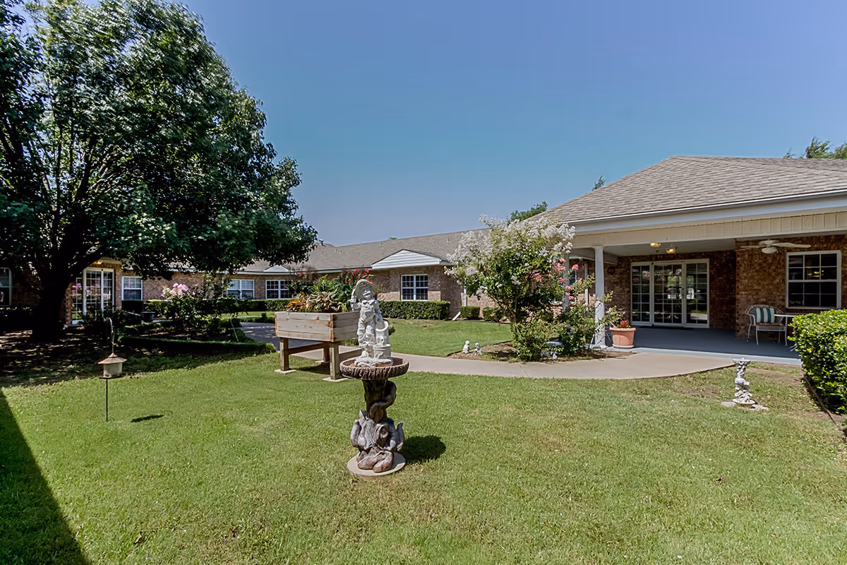 A grassy courtyard with a decorative fountain, planter boxes, and landscaping in front of a single-story senior living building with a covered porch.