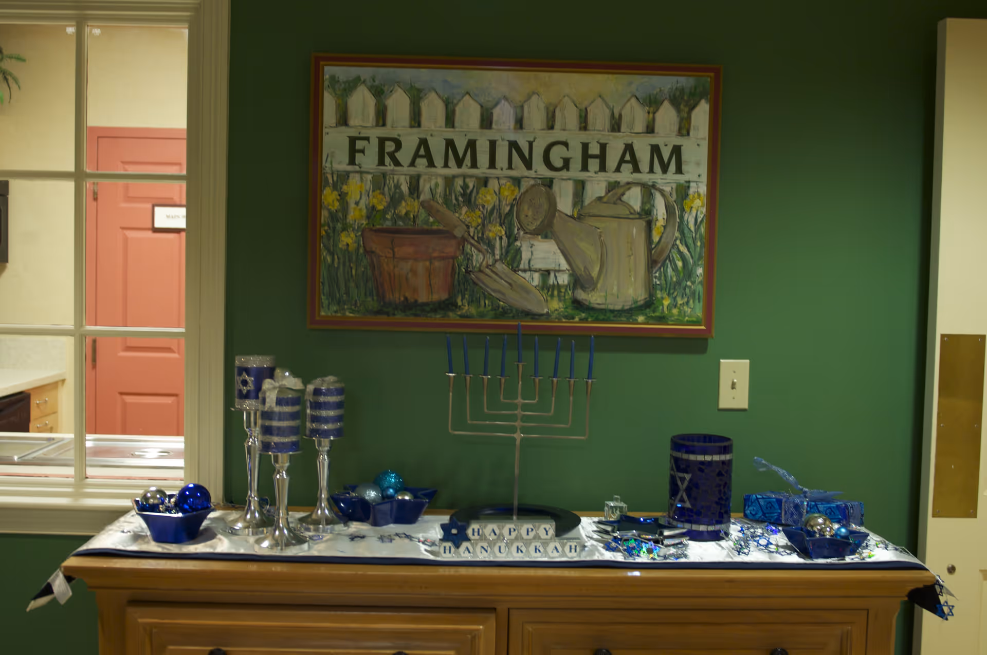 A wooden cabinet against a green wall decorated with Hanukkah items including blue and silver candles, a menorah with blue candles, blue decorative bowls with ornaments, and a 'Happy Hanukkah' sign. Above the cabinet is a framed painting of a white picket fence with gardening tools and the word 'FRAMINGHAM'. A window and a pink door are visible to the left.
