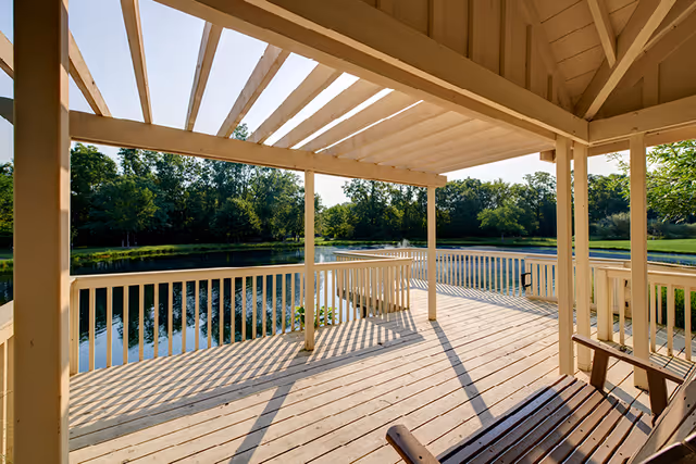A wooden deck with a pergola-style roof overlooking a calm pond surrounded by trees and greenery on a sunny day. The deck has white railings and a wooden bench in the foreground.