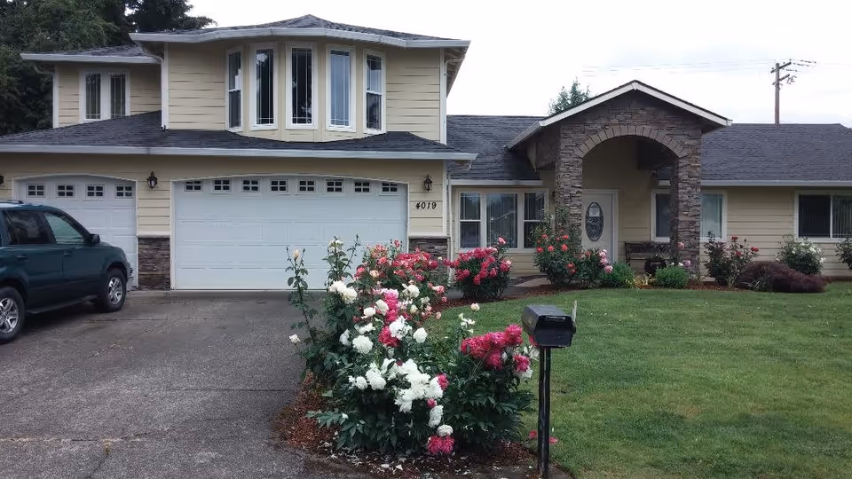 Front exterior view of a two-story residential-style building with beige siding and stone accents, featuring a three-car garage, a driveway with a parked green SUV, a well-maintained lawn, and flower beds with pink and white blooming flowers. A black mailbox is visible near the driveway.