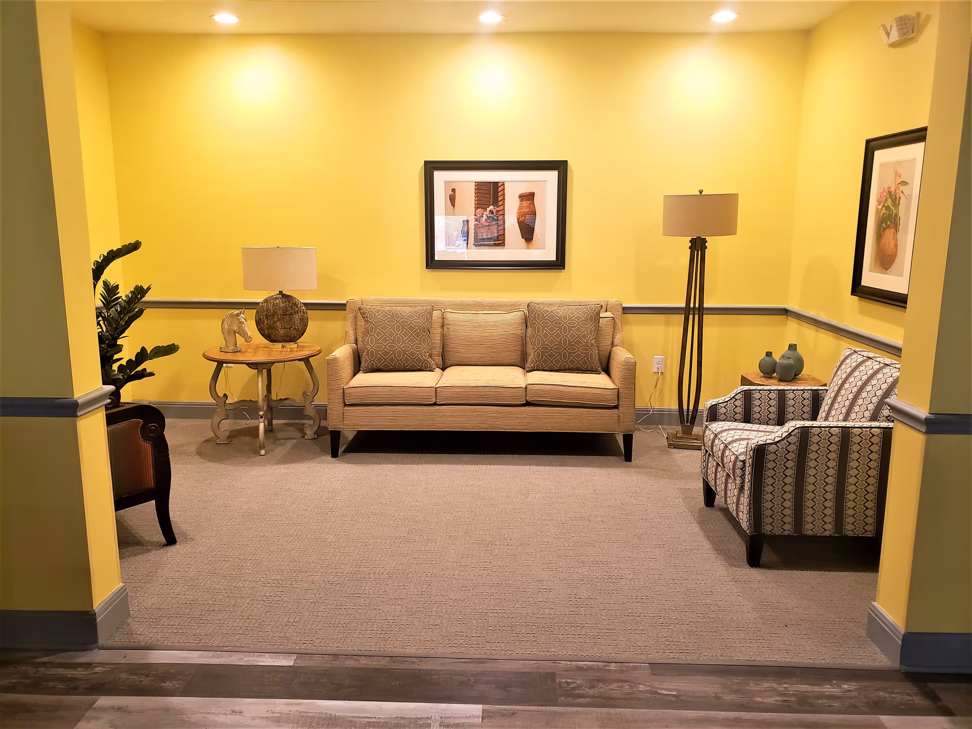 A cozy sitting area with yellow walls featuring a beige sofa with two patterned cushions, a patterned armchair, a wooden side table with a lamp and decorative horse head, a tall floor lamp, two framed pictures on the walls, and a potted plant in the corner.