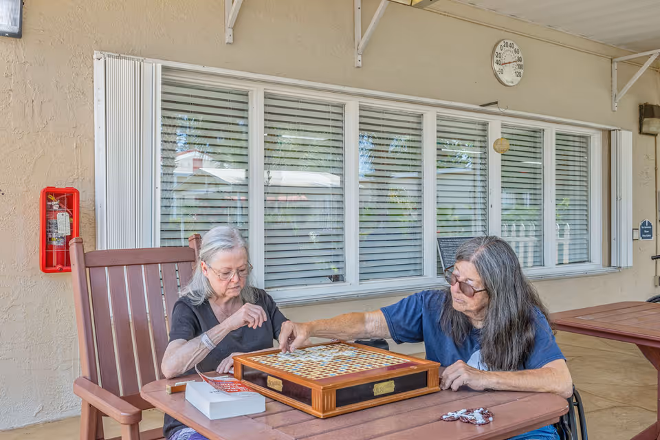 Two elderly women sitting at a wooden table outdoors playing a board game. One woman has gray hair and glasses, wearing a black shirt, and the other has long gray hair and glasses, wearing a blue shirt. Behind them is a beige wall with a window, a fire extinguisher, and a round thermometer.
