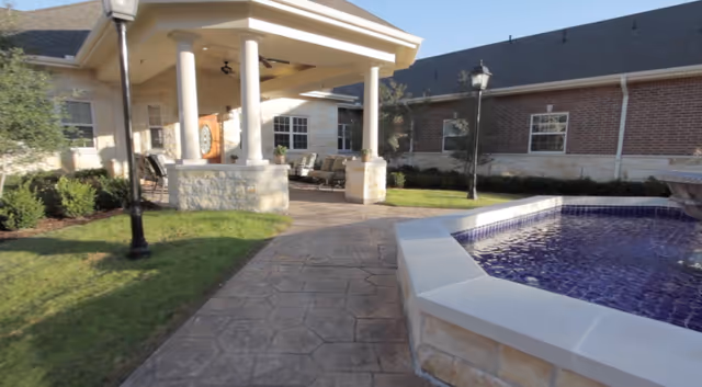 Outdoor patio area at a senior living facility with a covered seating area supported by white columns, a stone walkway, a decorative water feature with blue tiles, green grass, and a brick building in the background.
