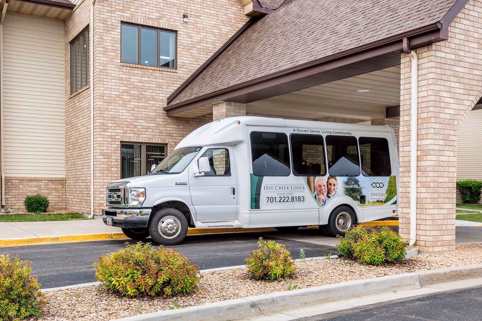 A white shuttle van parked under a covered entrance of a brick building. The van has signage for Hay Creek Lodge Senior Living with a phone number and images of smiling elderly people and a caregiver. There are small bushes and landscaping in front of the van.