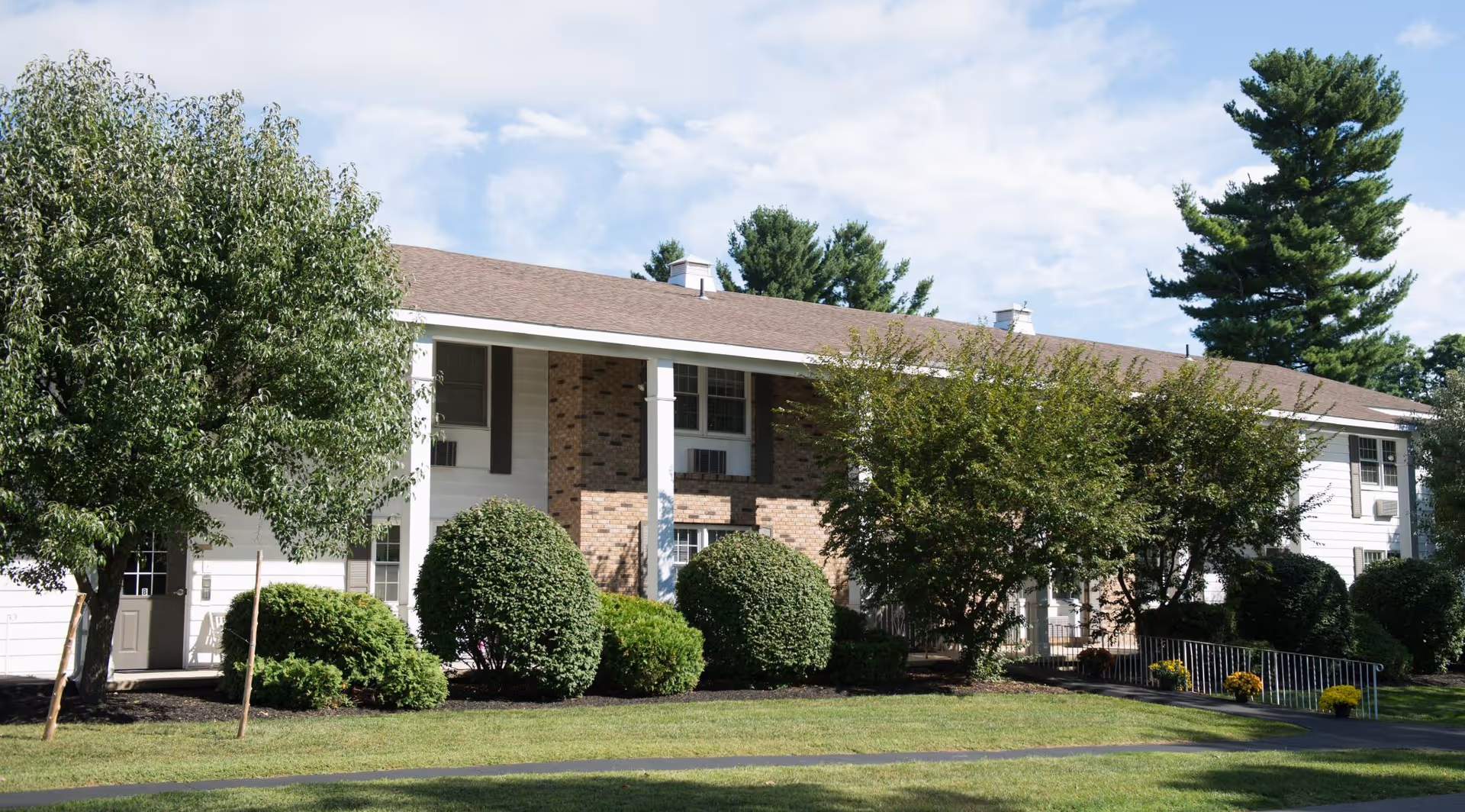 Exterior view of a two-story residential building with white siding and brick accents, surrounded by green trees and bushes under a partly cloudy sky.