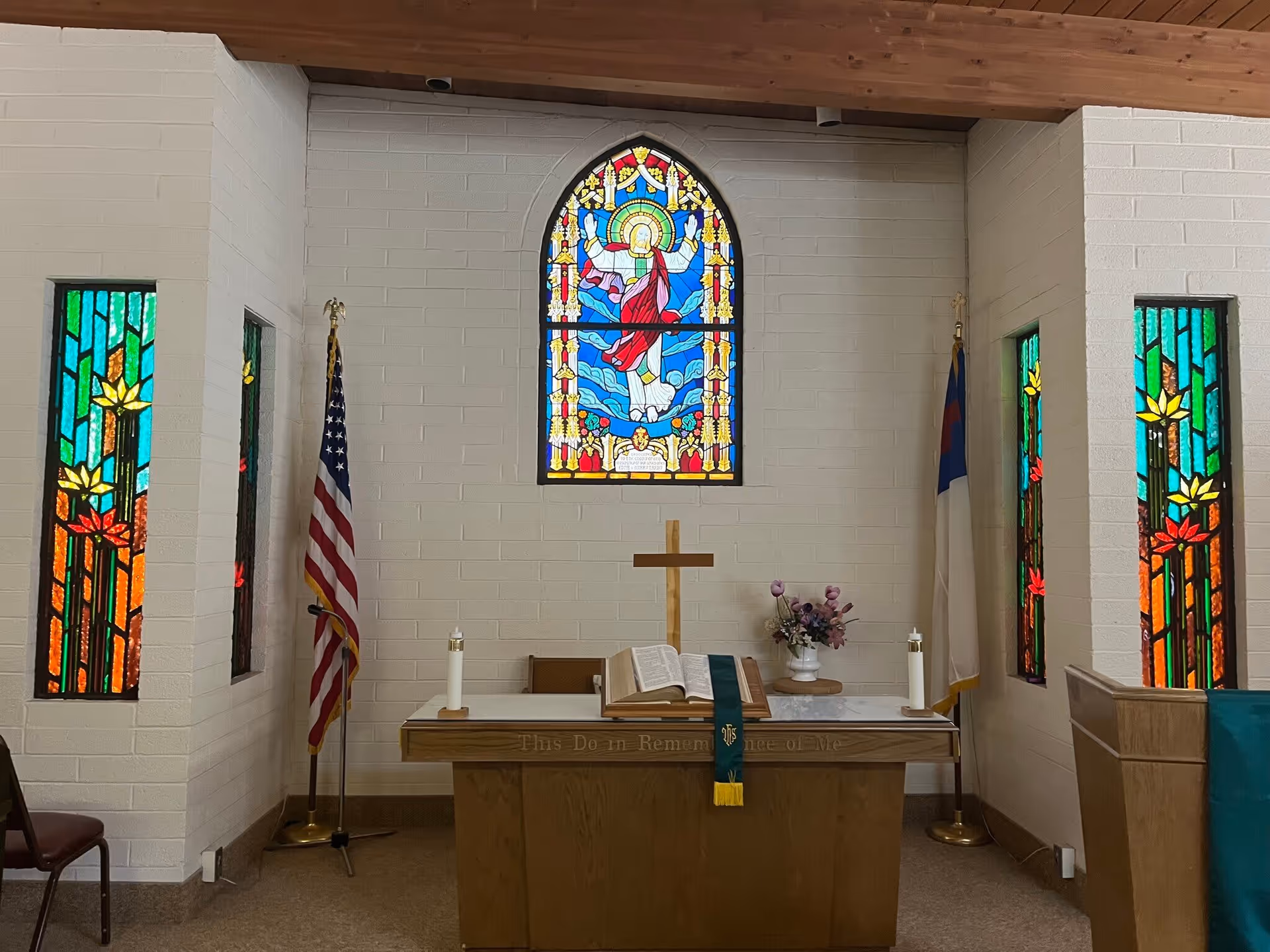 Chapel interior with an altar, wooden cross, open Bible, candles, flags, and colorful stained glass windows.