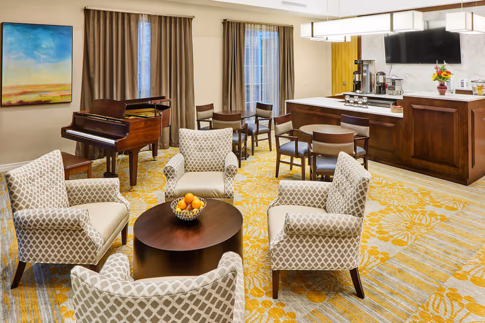 A cozy living room area with four patterned armchairs arranged around a round wooden coffee table holding a bowl of fruit. In the background, there is a grand piano near two windows with brown curtains, a small dining area with tables and chairs, and a wooden counter with a coffee station and a flat-screen TV mounted on the wall above it. The room features a yellow and gray floral carpet and a painting on the wall.