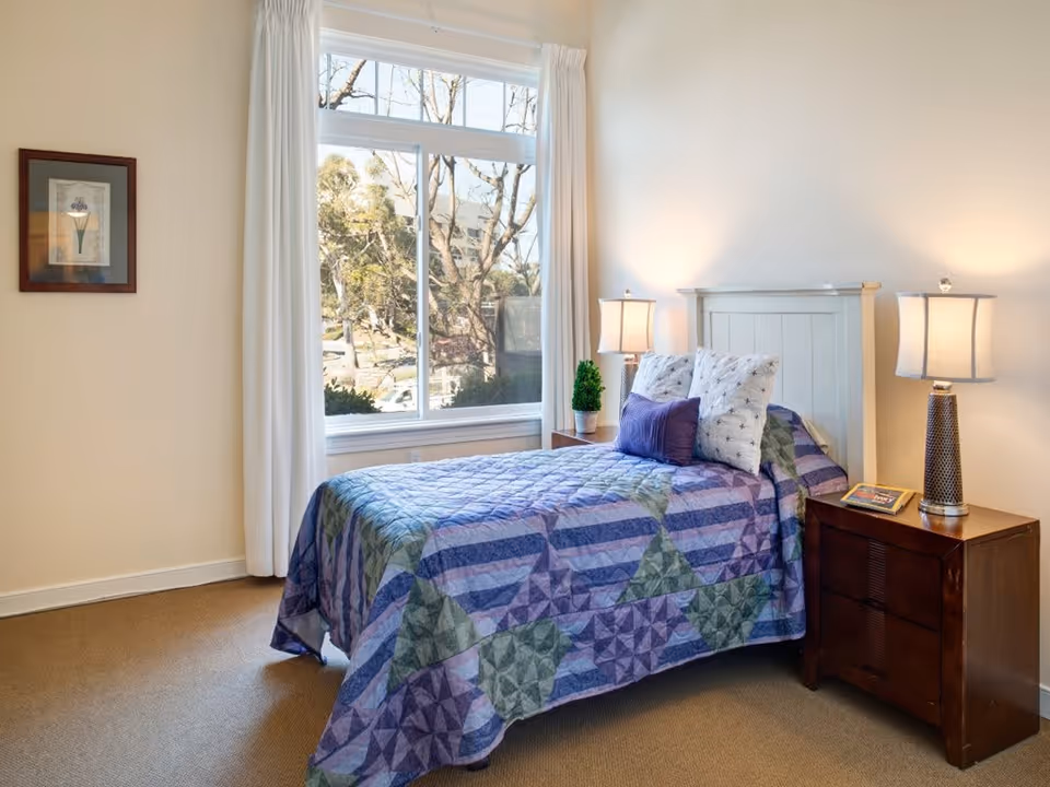 A cozy bedroom with a single bed covered in a colorful quilt featuring purple, green, and blue patterns. The bed has three pillows, two white with star patterns and one solid purple. There are two wooden nightstands on either side of the bed, each with a lamp. A small potted plant and a book are on the left nightstand. A large window with white curtains lets in natural light and shows trees outside. A framed floral artwork hangs on the wall to the left.